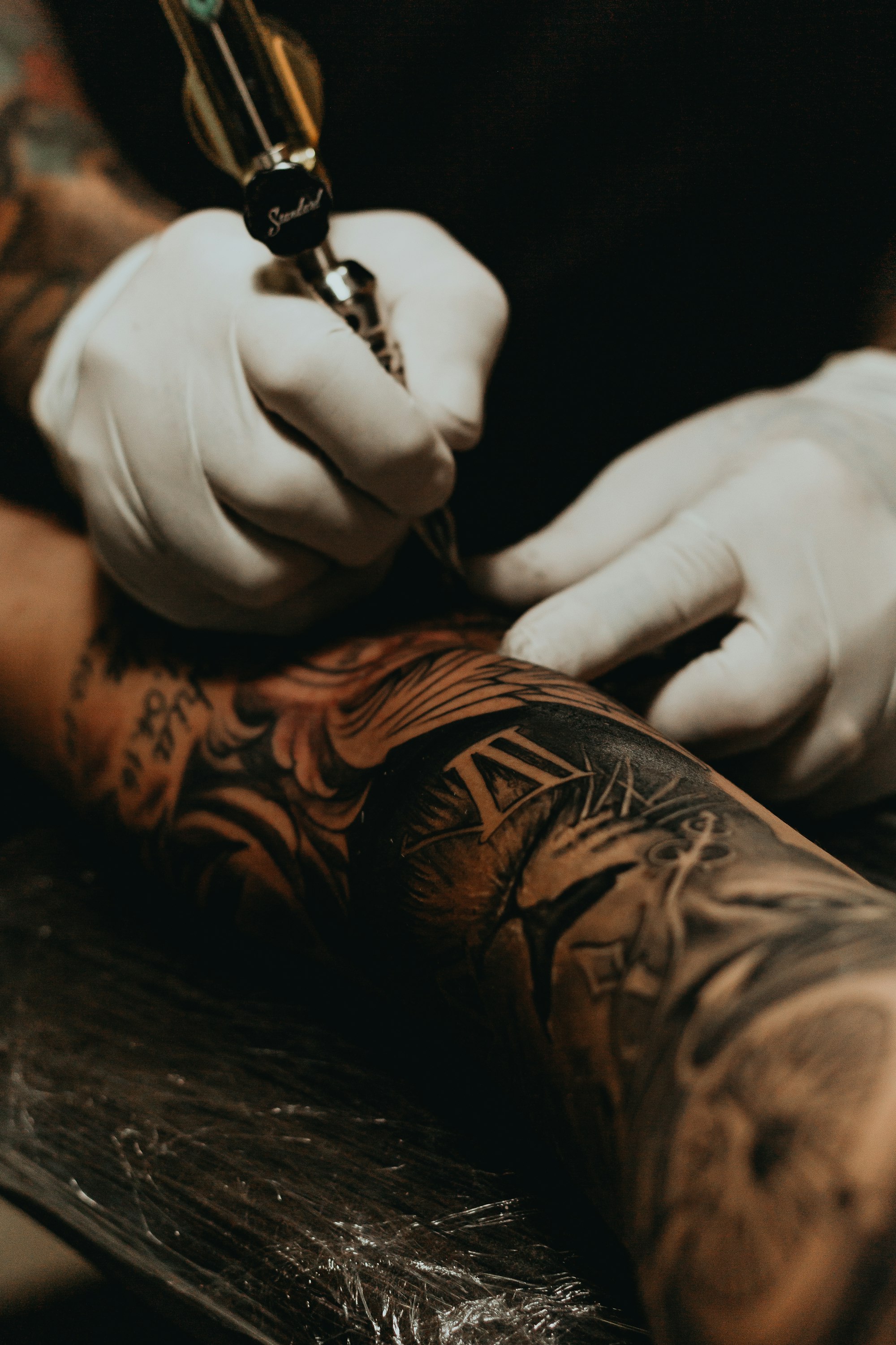 Gloved tattoo artist working on a heavily tattooed forearm, close up on the machine and fresh linework