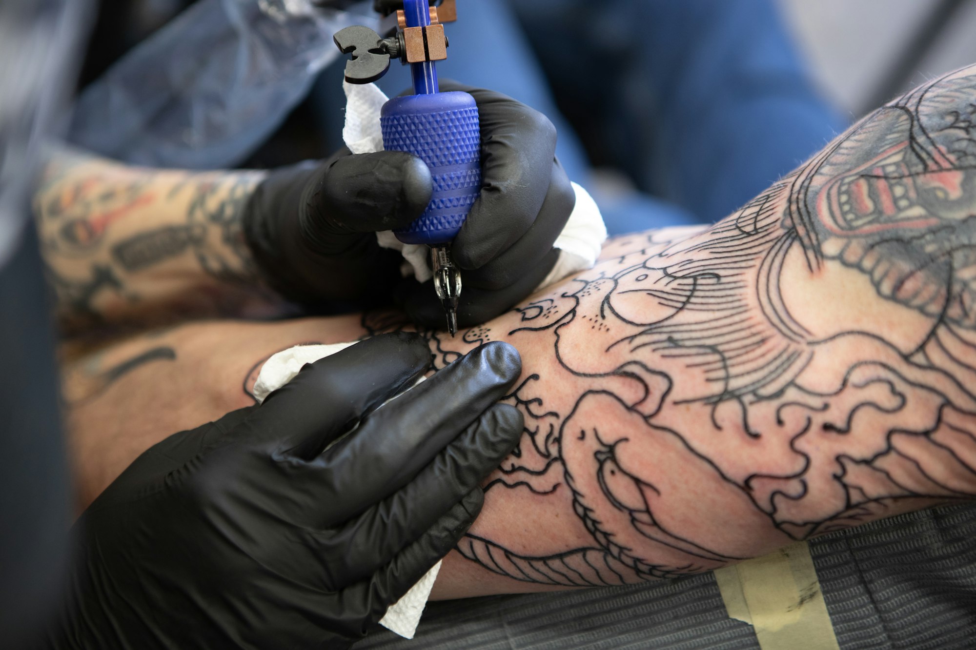Tattoo artist in black gloves applying fine linework to a client’s arm with a rotary machine