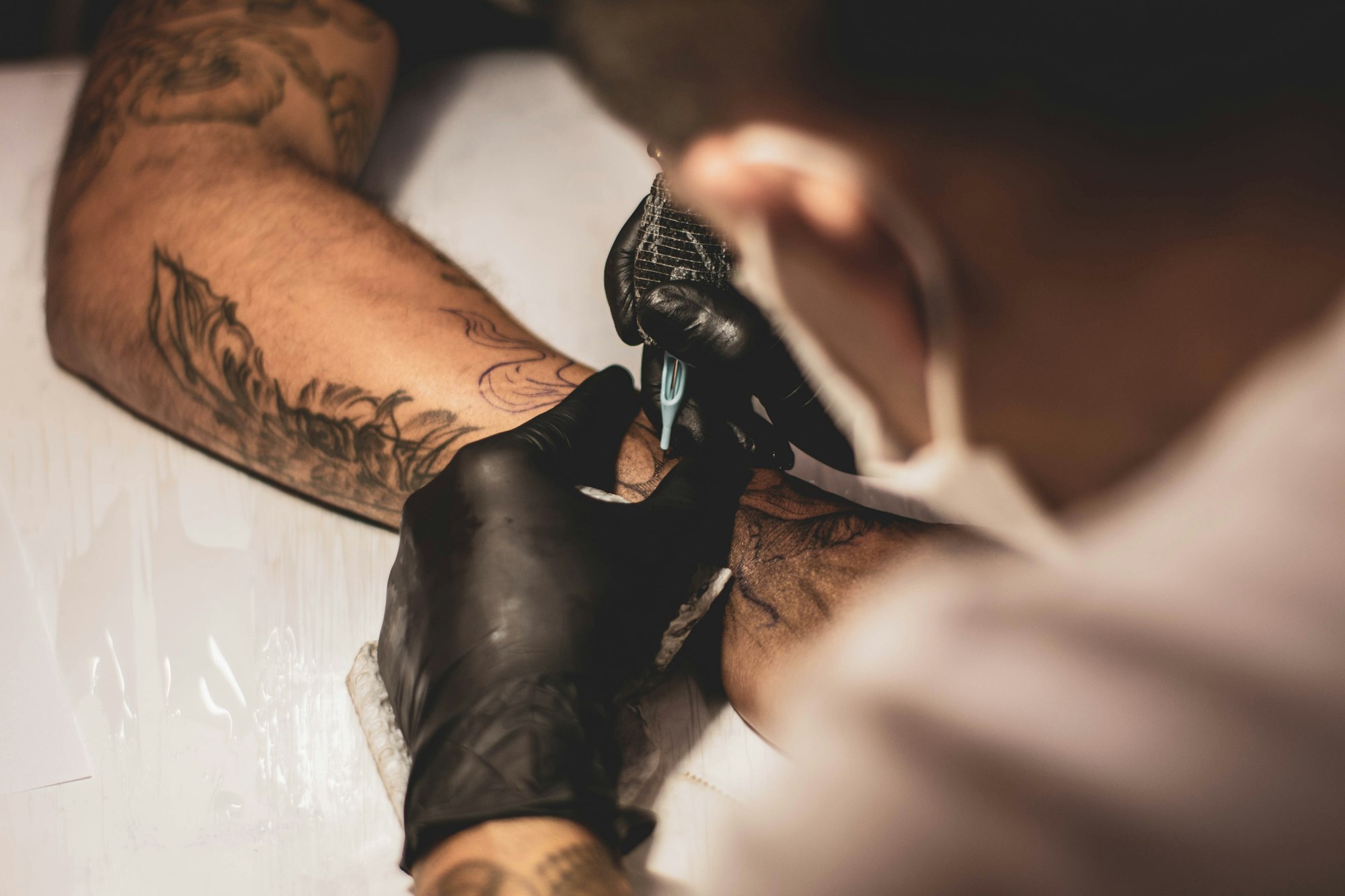 Shallow-focus photo of an artist tattooing a client’s right arm, machine and hand in sharp detail